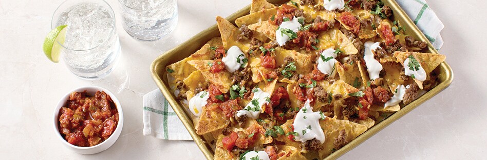 Sheet pan of nachos topped with seasoned beef, shredded cheese, sour cream and salsa.