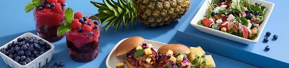 Spread of summer fruit, including blueberries, whole pineapple and a frozen berry drink with a berry salad and a pineapple pulled pork barbecue sandwich.