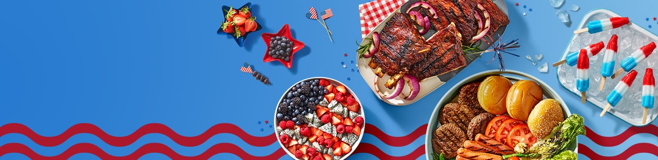 Festive 4th themed table showing grilled ribs, a bowl filled with fresh berries, firecracker popsicles and a platter of grilling essentials like buns, tomatoes, burgers, hot dogs and lettuce. 