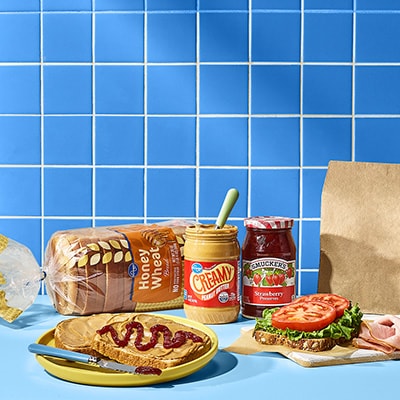 A kitchen counter with a loaf of honey wheat bread, open jars of peanut butter and strawberry jam, and a yellow plate holding two slices of bread—one with PB&J, the other with lettuce and tomato. Deli meat and a brown paper bag are nearby.