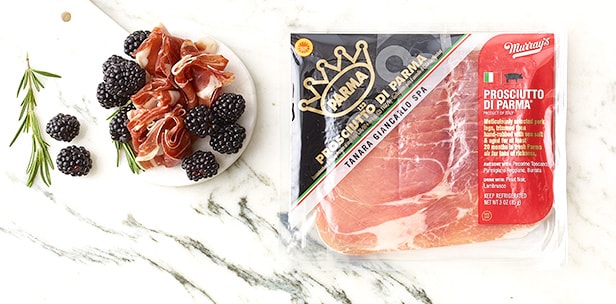 Plate of prosciutto and blackberries next to a package of Murray’s prosciutto on a white marble surface.