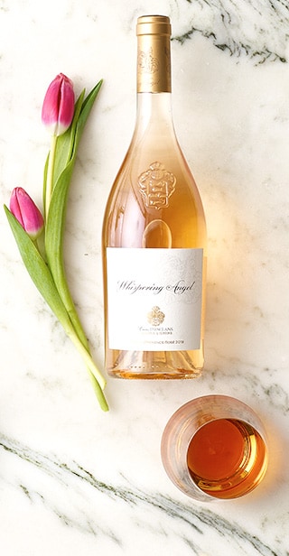 Bottle and glass of Chateau D’Esclans Whispering Angel Rose wine and pink tulips on a white marble surface.