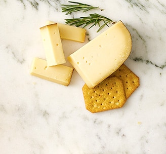 Block and slices of Murray’s Estate Gouda cheese, crackers, and a sprig of rosemary on a white marble surface.