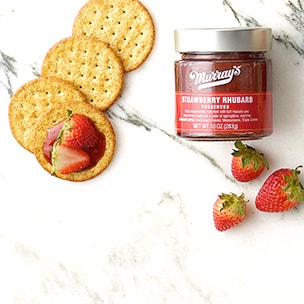 Jar of Murray’s Strawberry Rhubarb Preserves, crackers and strawberries on a white marble surface.