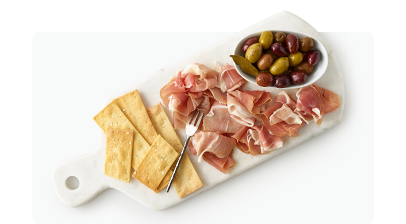 White stone charcuterie platter on top of white surface with crackers, prosciutto and a bowl of olives