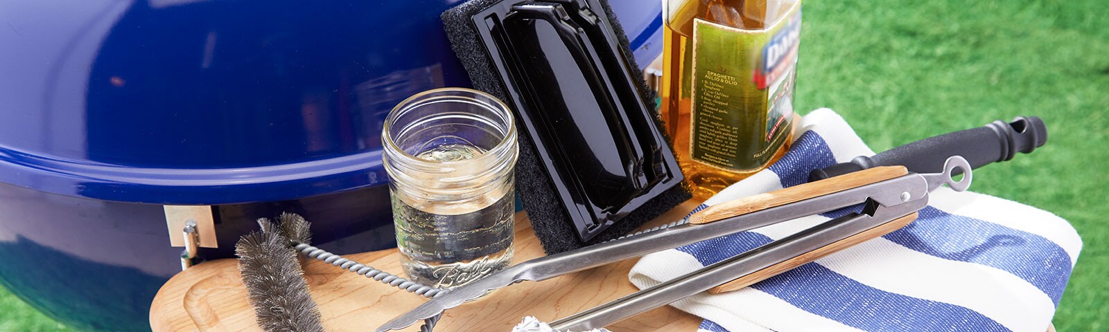 Blue outdoor drill with an attached shelf, on which sits a cloth, cooking oil, tongs, a wire brush, tin foil, scrub brush and mason jar. Blue outdoor drill with an attached shelf, on which sits a cloth, cooking oil, tongs, a wire brush, tin foil, scrub brush and mason jar.