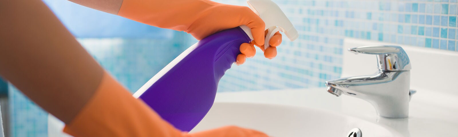 Two arms wearing rubber gloves cleaning a bathroom sink, spray bottle in the left hand with a cloth to wipe in the right Two arms wearing rubber gloves cleaning a bathroom sink, spray bottle in the left hand with a cloth to wipe in the right