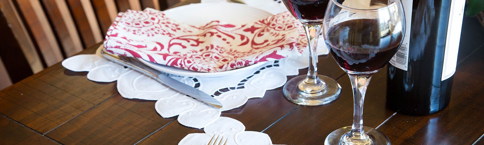 A wooden table, atop sits two wine glasses filled with red wine and two place settings