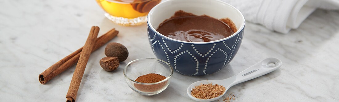 On a marble countertop nutmeg, cinnamon, cinnamon sticks and a container of honey sit surrounding a bowl of them mixed together