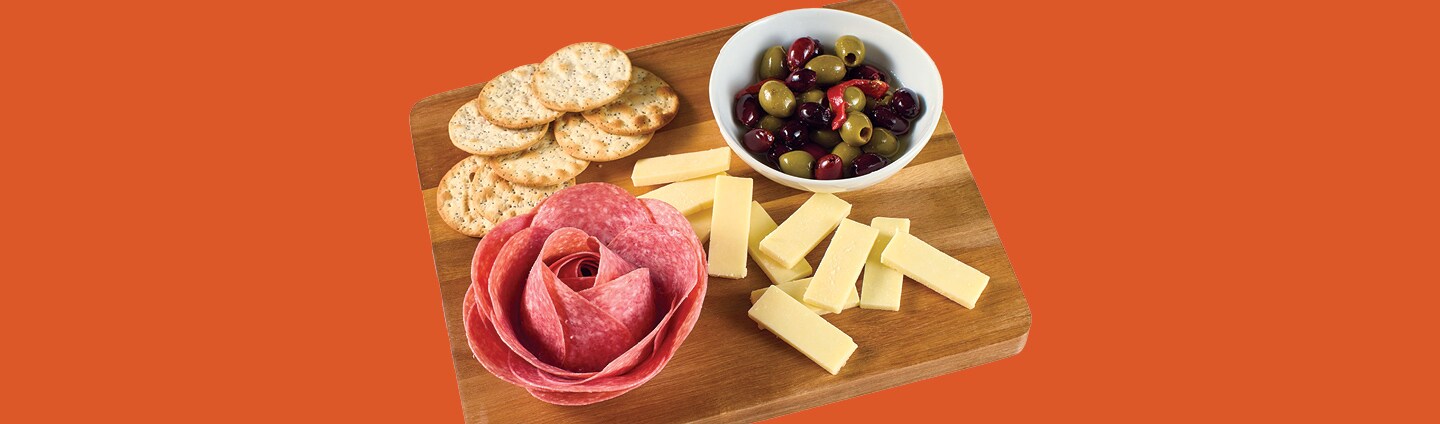 Salami rose on a charcuterie board with cheese slices, crackers and bowl of olives, floating on an orange background