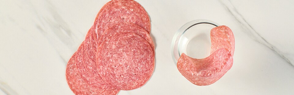 Salami slices layered over wine glass rim, on a white marble surface.