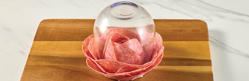 Wine glass flipped upside down with salami slices layered underneath in a rose shape. Wine glass is on a wooden cutting board, on a white marble surface.