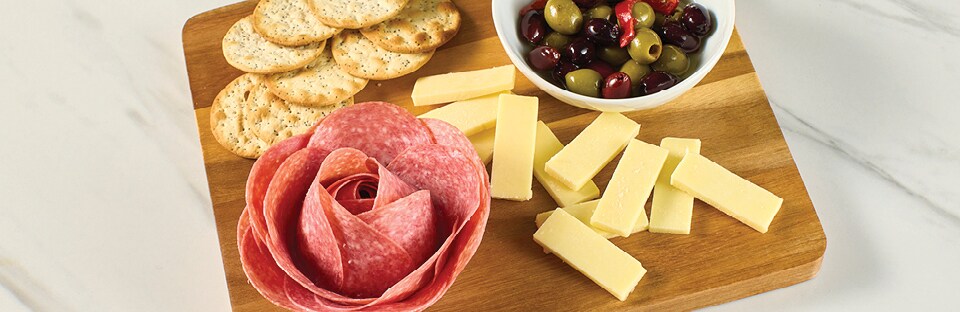 Salami rose on a cutting board with cheese slices, crackers and bowl of olives, on a white marble surface