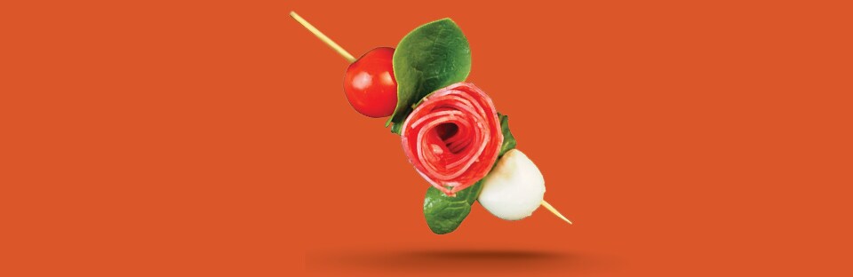 A skewer showing the Single “Stem” Method, with a cherry tomato, basil leaf, salami rose, a second basil leaf and a mini mozzarella ball. Single skewer is floating on an orange background
